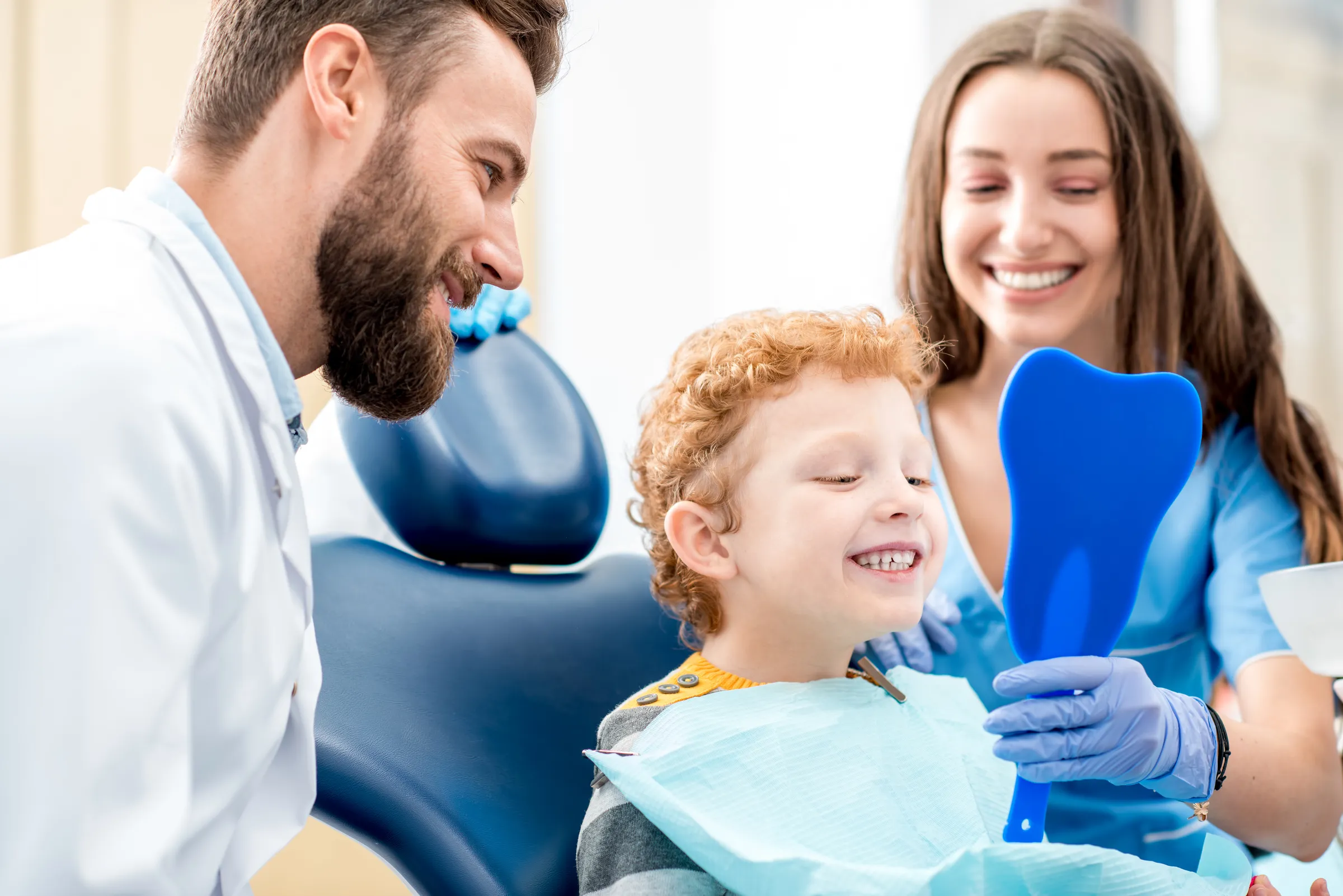 Boy in a dental chair smiling at a mirror with his mother and dentist