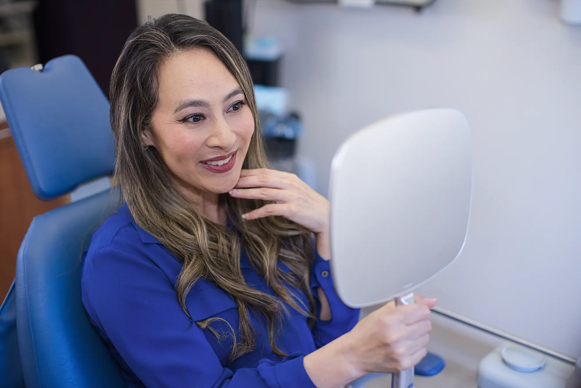 Smiling patient in the dental chair holding a hand mirror and touching her chin