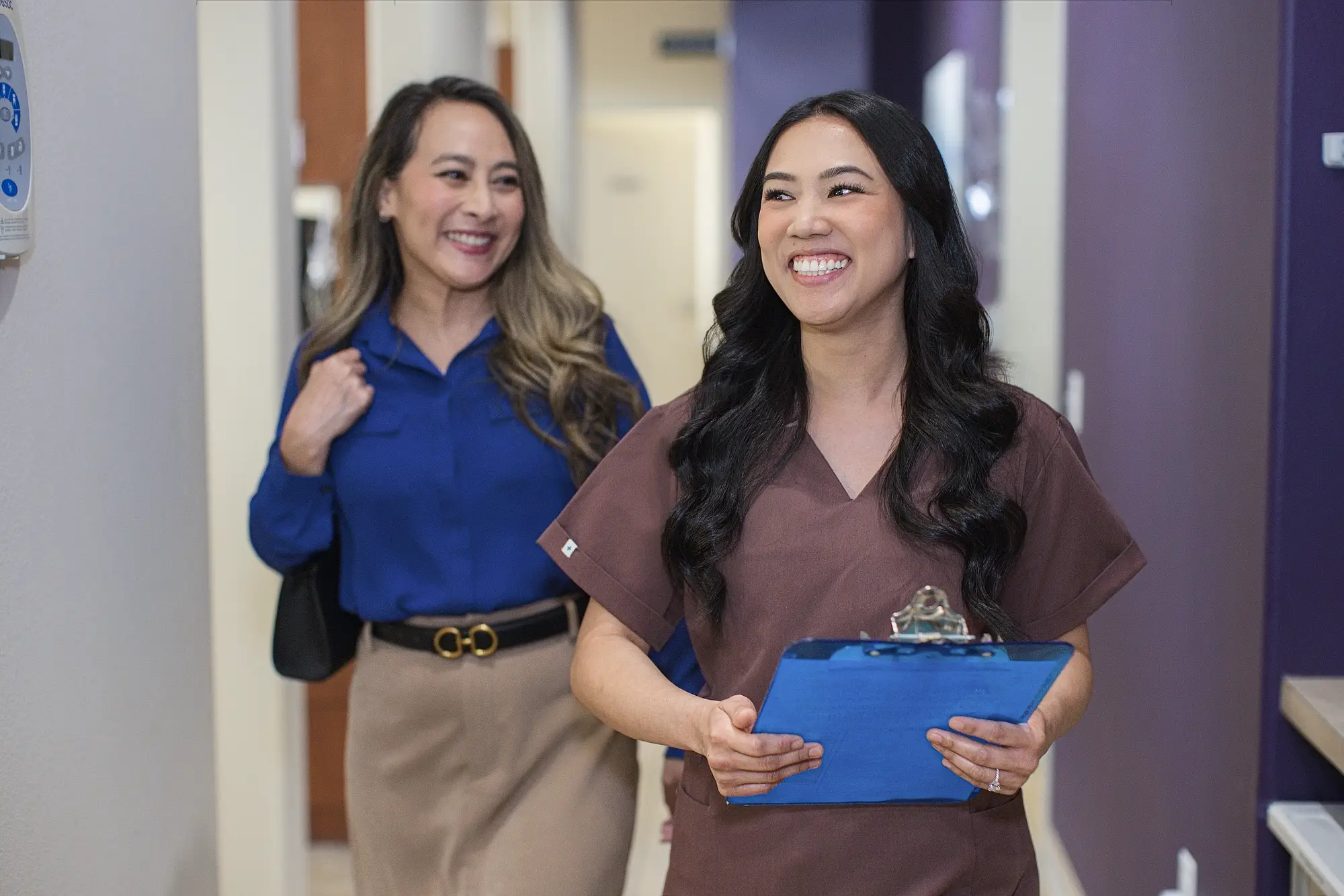 Team member in brown scrubs walking a smiling patient down the hallway
