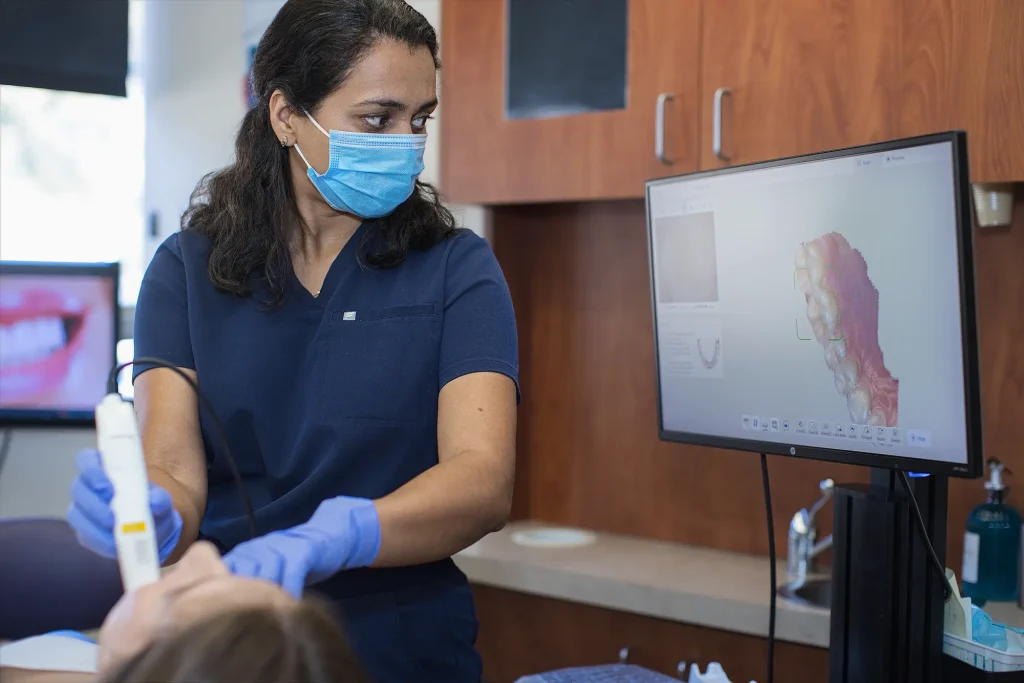 Dental team member in a mask holding an intraoral scanner beside a monitor showing a 3D model of teeth